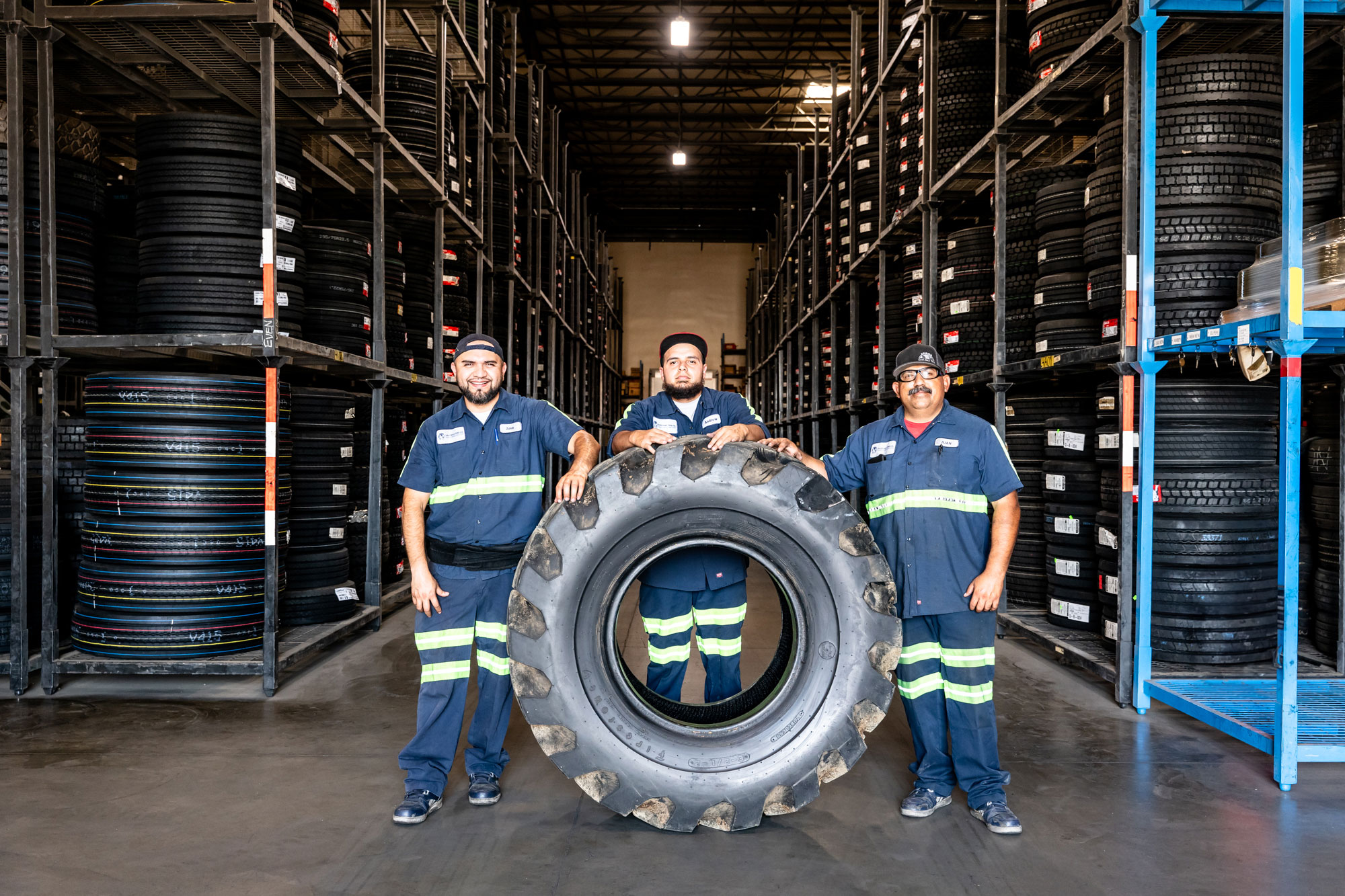 Empleados de East Bay Tire de pie con un neumático grande en el pasillo de un almacén.